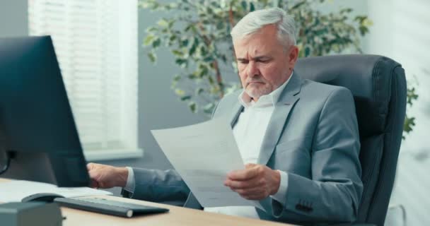 Un experimentado hombre de negocios, de cabello gris jefe de empresa está sentado en el escritorio en una cómoda silla del presidente, mirando los documentos traídos a él, leyéndolos cuidadosamente, preguntando a un colega — Vídeos de Stock