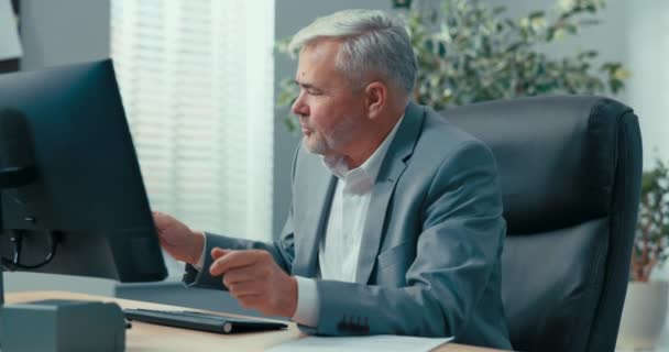 Un experimentado hombre de negocios, de cabello gris jefe de empresa está sentado en el escritorio en una cómoda silla del presidente, mirando los documentos traídos a él, leyéndolos cuidadosamente, preguntando a un colega — Vídeos de Stock