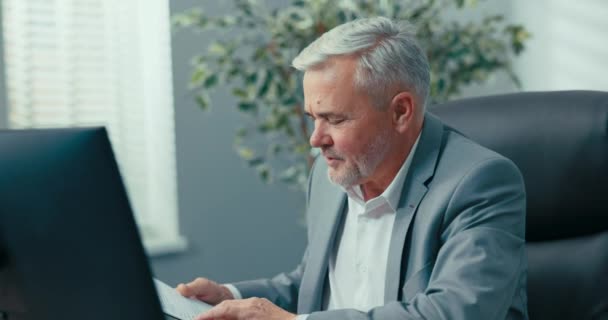 A cégvezető, ősz hajú, elegáns dzsekit viselő idős férfi, videokamera segítségével távolsági konferenciát tart az interneten keresztül, dokumentumokat, statisztikákat és eredményeket mutat be. — Stock videók