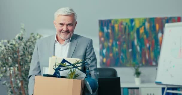Вихід на пенсію досвідченого давнього директора, менеджера, керівника компанії, літнього чоловіка з сірим волоссям тримає картонну коробку з упакованими речами в руках, дивиться всередину, посміхається, відпочиває, виходить на пенсію — стокове відео