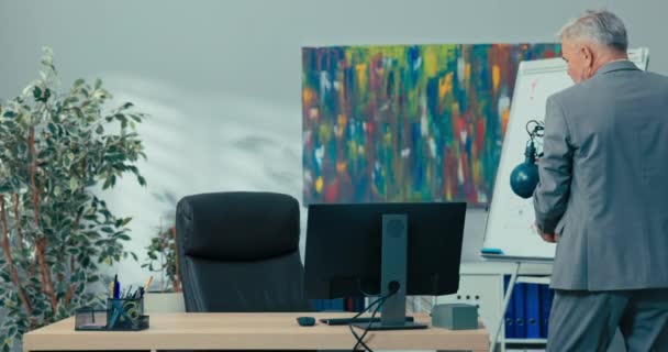 Амбіційний літній чоловік з сірим волоссям у сорочці стоїть перед столом у корпоративному офісі, тримаючи в руках упаковані речі, приходить до корпорації, щоб бути підвищений до нової роботи розпакувальні аксесуари — стокове відео