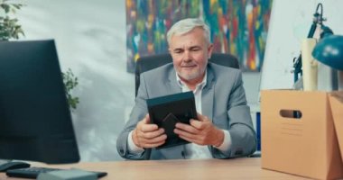 Yeni çalışan, gri saçlı, gömlekli yaşlı bir adam ofisinde yanında paketlenmiş eşyalarla dolu bir kutuda oturuyor. Tezgahın yanındaki resim çerçevesini kaldırıyor. Çalışma yerinin yanındaki bilgisayar hazırlıyor.