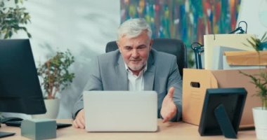 Gri saçlı zarif adam ofisten uzaktan konferans düzenliyor, dizüstü bilgisayarın önünde oturuyor, meslektaşlarına el sallıyor, web kamerasında online iş görüşmeleri yapıyor, görüntülü sohbet yapıyor.