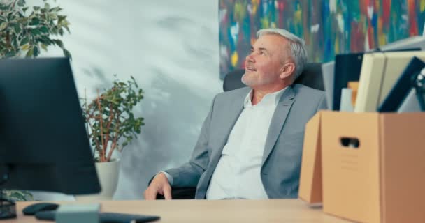 Lächelnde leitende Angestellte des Unternehmens sitzen im bequemen Drehstuhl neben dem Computer und schauen sich um, wie der elegante Mann mit den grauen Haaren seinen letzten Arbeitstag vor der Rente verbringt — Stockvideo
