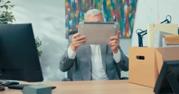 Новий корпоративний працівник старший чоловік з сірим волоссям у сорочці сидить за столом в офісі на стільниці лежить картонна коробка з аксесуарами бізнесмен витягує ноутбук і відкриває його готується до роботи — стокове відео