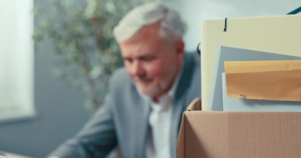 Shot from behind cardboard box with employees belongings, workstation equipment documents concentrated elderly man with gray hair looking at laptop screen reports getting into work reading emails — Stock Video