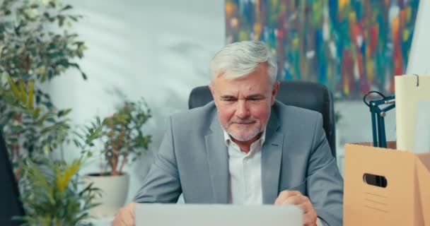 Gri saçlı takım elbiseli adam, uzaktan kumandalı bir konuşmayı laptopun önünde oturarak web kamerası görüntülü sohbet üzerinden iş arkadaşlarına el sallayarak el sallıyor. — Stok video