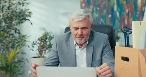 Hombre de traje con pelo gris lleva conversación remota desde la oficina sentado en el escritorio delante de la computadora portátil saludando a sus colegas reuniones en línea a través de webcam chat de vídeo hablar de impresiones gesticula — Vídeo de stock