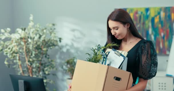 Nieuwe werknemer van het bedrijf staat in het midden van het kantoor naast het bureau met een kartonnen doos in haar handen met verpakte accessoires, apparatuur, kijkt naar binnen glimlacht acceptatie aan het werk, promotie — Stockvideo