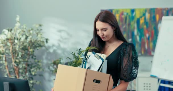 New employee of the company stands in middle of the office next to the desk holding a cardboard box in her hands with packed accessories, equipment, looks inside smiles acceptance to work, promotion — Stock Video