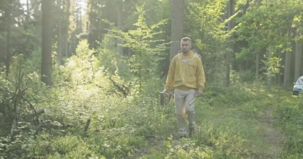 Mladý muž ve žluté bundě s kloboukem, který má záchrannou síť, míří lesem k včelínovi s úly, které včelař drží v náručí. — Stock video