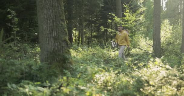 Un joven vistiendo una chaqueta amarilla con un sombrero que tiene una red de seguridad se dirige a través del bosque hacia un colmenar con colmenas un apicultor está sosteniendo accesorios — Vídeos de Stock