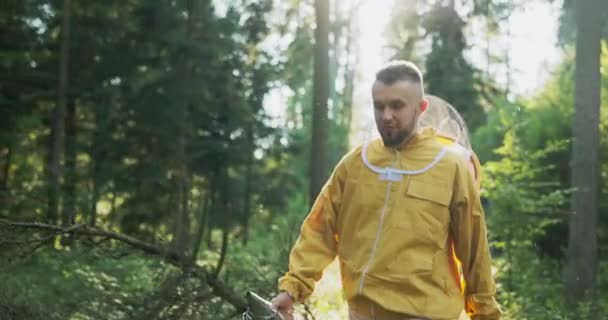 Un giovane uomo che indossa una giacca gialla con un cappello che ha una rete protettiva si sta dirigendo attraverso il bosco verso un apiario con orticaria, un apicoltore sta lavorando, si prende cura delle api — Video Stock