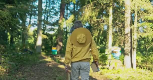 Imker steuert Bienenstöcke mit Utensilien in der Hand an, stapft durch den Wald zum Bienenstand, macht sich an die Arbeit — Stockvideo