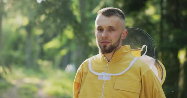A young bee keeper working in the woods by the apiary hives stands with the bite protection net removed from his head, an experienced beekeeping guy — Stock Video