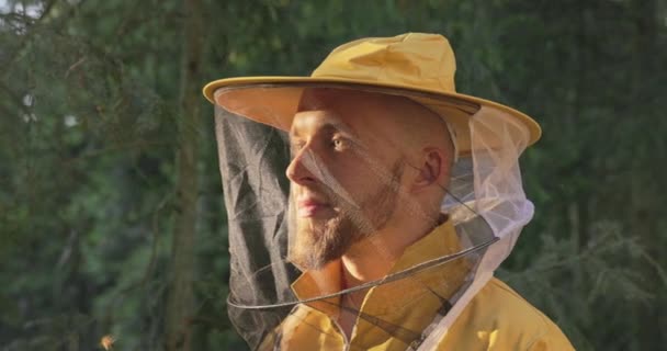 Sorridente apicoltore maschio nella foresta al mattino con il sole che sorge, indossando una tuta gialla faccia a rete per proteggere contro le punture di insetti, il ragazzo si prende cura delle api — Video Stock