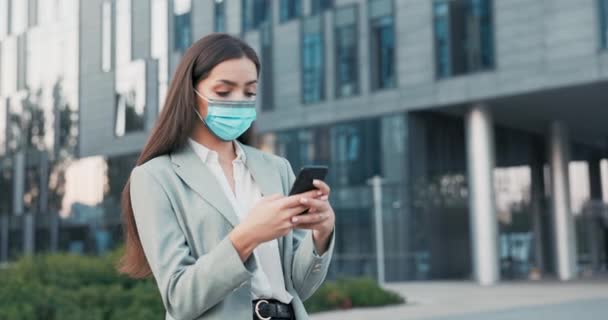 Uma mulher bonita vestida com uma camisa e jaqueta com máscara protetora no rosto está de pé na frente do prédio corporativo, quebrar do trabalho, segurando um telefone nas mãos, respondendo mensagens, e-mail — Vídeo de Stock