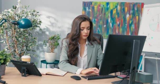 Attractive pretty brunette with dark eyes in jacket sits in comfortable chair in front of laptop, sexy boss secretary, beautiful manager, company owner, businesswoman, successful woman — стоковое видео