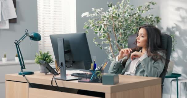 Женщина с длинными волосами классический макияж носить куртку сидит за столом в офисе опираясь на спинку стула обдумывая решение ситуации писать ответ клиенту во время нажатия на клавиатуру компьютера — стоковое видео