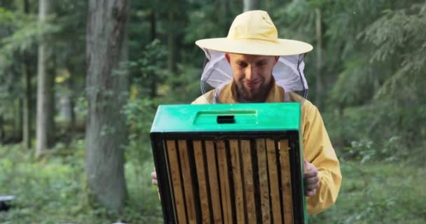 Молодой опытный улыбающийся человек, занимающийся пчеловодством, держит в руках пасеку с пчелами, смотрит на их работу в защитном костюме с сеткой для комаров на лице, чтобы защитить от укусов — стоковое видео