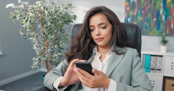 Seorang pekerja kantor yang santai telah istirahat dari pekerjaan, gadis cantik berpakaian elegan duduk nyaman di kursi di meja, memegang telepon di tangan, browsing media sosial, situs web, menulis pesan — Stok Video