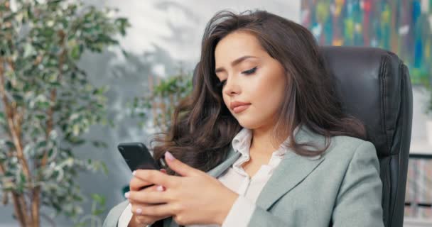 Un trabajador de oficina relajado tiene descanso del trabajo, una chica joven y bonita elegantemente vestida se sienta cómodamente en un sillón en el escritorio, sosteniendo un teléfono en la mano, navegando por las redes sociales, sitios web, escribiendo mensajes — Vídeos de Stock