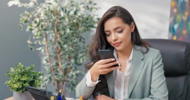 Sosyal medya, pazarlama ve müşteri iletişimi uzmanı şirket ofisinde telefon başında oturur, satış ilanları, reklamlar ekler, kameraya telefon tebessümleri koyar.