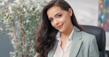 Gülümseyen, güzel, seksi, siyah gözlü, gülümseyen esmer, zarif giyimli kadın, şirket ofisinde oturan iş kadını, sekreter, asistan, kameraya göz kırpması.