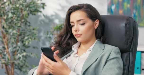 Un trabajador de oficina relajado tiene descanso del trabajo, una chica joven y bonita elegantemente vestida se sienta cómodamente en un sillón en el escritorio, sosteniendo un teléfono en la mano, navegando por las redes sociales, sitios web, escribiendo mensajes — Vídeos de Stock