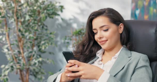 Um trabalhador de escritório sorridente tem uma pausa do trabalho, menina bonita elegantemente vestida senta-se confortavelmente em uma poltrona na mesa, segurando um telefone na mão, navegando nas mídias sociais, sites, escrevendo mensagens — Vídeo de Stock