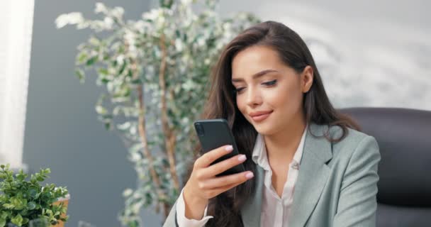 En leende kontorsarbetare har en paus från jobbet, söt ung flicka elegant klädd sitter bekvämt i en fåtölj vid skrivbordet, hålla en telefon i handen, bläddra sociala medier, webbplatser, skriva meddelanden — Stockvideo