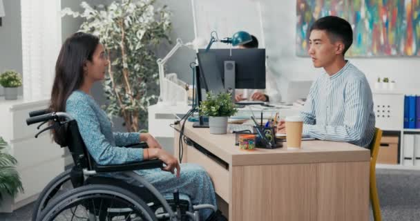 Jobbintervju med funktionshindrade kvinna i rullstol, sträng manlig chef för asiatisk-koreanska skönhet ställer frågor till flicka i klänning, de sitter på företagets kontor på två sidor av skrivbordet killen lyssnar noga för att svara — Stockvideo