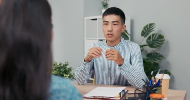 Job interview met gehandicapte vrouw in rolstoel, glimlachende baas man van Aziatische-Koreaanse schoonheid stelt vragen aan meisje in jurk ze zitten in het kantoor van het bedrijf aan twee zijden van het bureau man luistert zorgvuldig te beantwoorden — Stockvideo