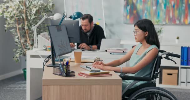 Workday en la oficina, corporación, empresa, compañeros de trabajo están trabajando en computadoras, redacción de documentos, mujer en gafas con belleza asiática está sentado en el escritorio en silla de ruedas, ella es discapacitada, después de un accidente — Vídeos de Stock