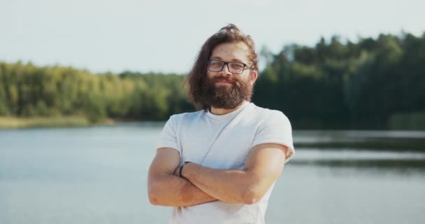 Et portræt af en mand med langt hår og skæg, briller på næsen, en dreng lægger armene krydsede, kigger ind i kameraet med et smil, tillid, i baggrunden en sø, sommer eftermiddag – Stock-video