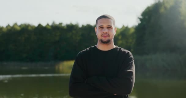 A man in a black sweatshirt is standing in background of a lake, he has his arms crossed over chest, he is looking into the camera with a serious face, a handsome guy is spending time outdoors — Stock Video