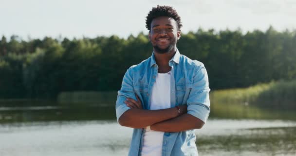 En stilig man med kort mörkt lockigt hår och skägg står mot en sjö med armarna över bröstet, leende sympatiskt — Stockvideo
