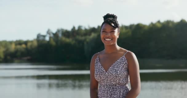 Beautiful smiling cheerful woman in floral dress is hanging out at the lake, her hair is tied up in a bun, she turns towards the camera, stunning look — Stock Video