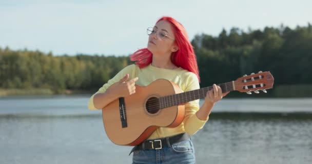 En glad flicka med rosa hår och glasögon står nära sjön, håller en gitarr i händerna, spelar och sjunger favoritlåtar, leker runt, dansar — Stockvideo