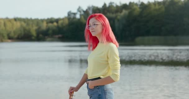 Vrolijk meisje met roze haar geblazen door de wind draagt een bril staat in de buurt van het meer, de hand rust op de gitaar, glimlachen naar de camera, losse outfit — Stockvideo