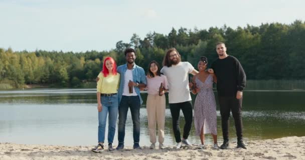 Portret sześciu znajomych bawiących się razem nad jeziorem na plaży, luźno ubranych spędzających wieczór, świętujących spotkanie, pijących piwo z szklanych butelek, grupa studentów — Wideo stockowe