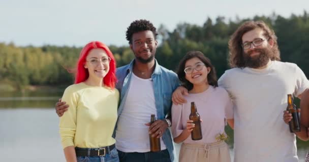 Un gruppo di studenti di diverse nazionalità si incontrano sul lago durante le vacanze estive, si abbracciano in piedi sulla spiaggia bere birra da bottiglie di vetro festeggiare esami di passaggio divertirsi insieme — Video Stock