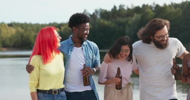 Grupa uczniów różnych narodowości spotyka się na jeziorze podczas wakacji letnich, przytulają się stojąc na plaży pijąc piwo z szklanych butelek świętując zdanie egzaminów baw się dobrze razem — Wideo stockowe