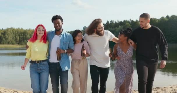 Группа студентов разных национальностей встречается на отдыхе на озере, они обнимают друг друга, идут к камере, смеются, дурачатся, держа в руках пивные бутылки. — стоковое видео