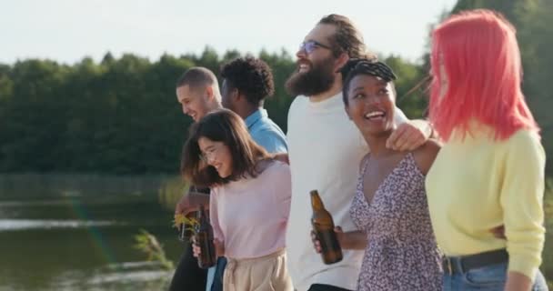 Φίλοι συναντιούνται στη λίμνη για να περάσουν χρόνο μαζί, αγκαλιάζονται περπατώντας μπροστά, γελώντας, χαζεύοντας κρατώντας μπουκάλια μπύρας στα χέρια τους, τα μαλλιά τους ανεμισμένα από τον άνεμο — Αρχείο Βίντεο