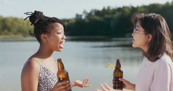 Dos hermosos y sonrientes amigos pasan tiempo juntos en la orilla del lago, sosteniendo botellas de cerveza de vidrio en sus manos, tonteando, riendo, contando una historia abrazándose con alegría — Vídeos de Stock