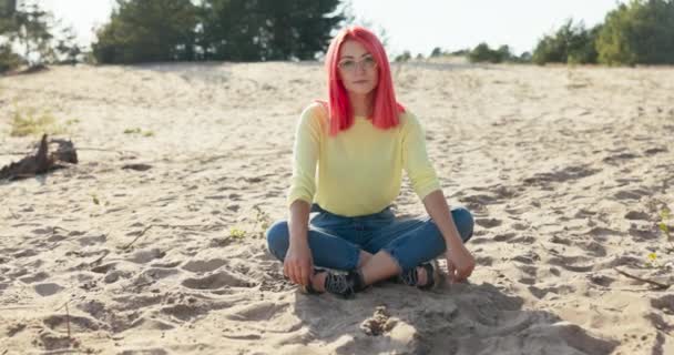 En smuk smilende pige sidder på sand udenfor på stranden, lyserødt hår blæses væk af vinden, hun blæser det ud af ansigtet, hun har briller på næsen, hun ser ind i kameraet. – Stock-video