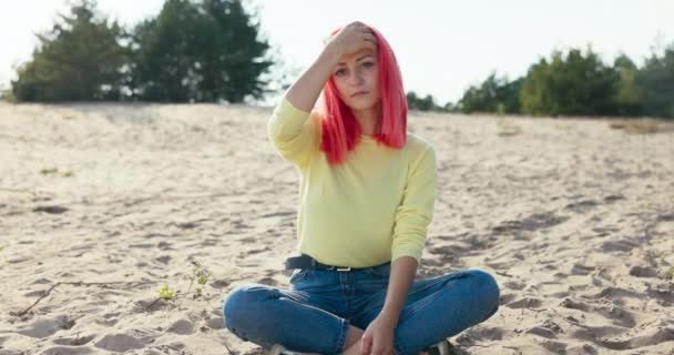 En smuk seriøs pige sidder på sandet udenfor på stranden, lyserødt hår blæses væk af vinden, hun blæser det ud af hendes ansigt, dybe øjne, fregner på ansigtet. – Stock-video