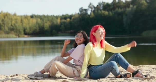 Meninas sorridentes de óculos estão sentadas na areia da praia perto do lago, encostadas umas às outras, mulheres bonitas estão rindo, brincando, passando tempo juntas — Vídeo de Stock