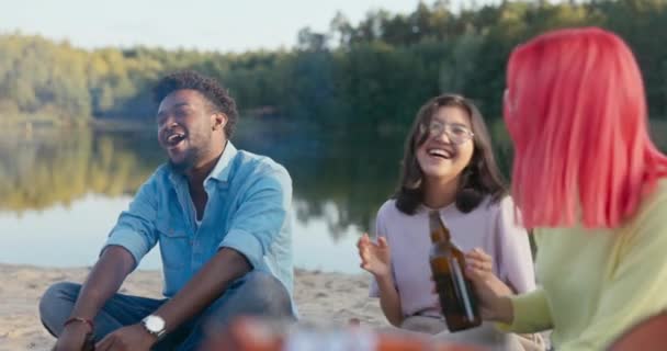 En grupp vänner sitter på stranden vid sjön, dricker öl vid brasan, sjunger sånger, svänger från sida till sida till musikens rytm, människor av olika nationaliteter — Stockvideo